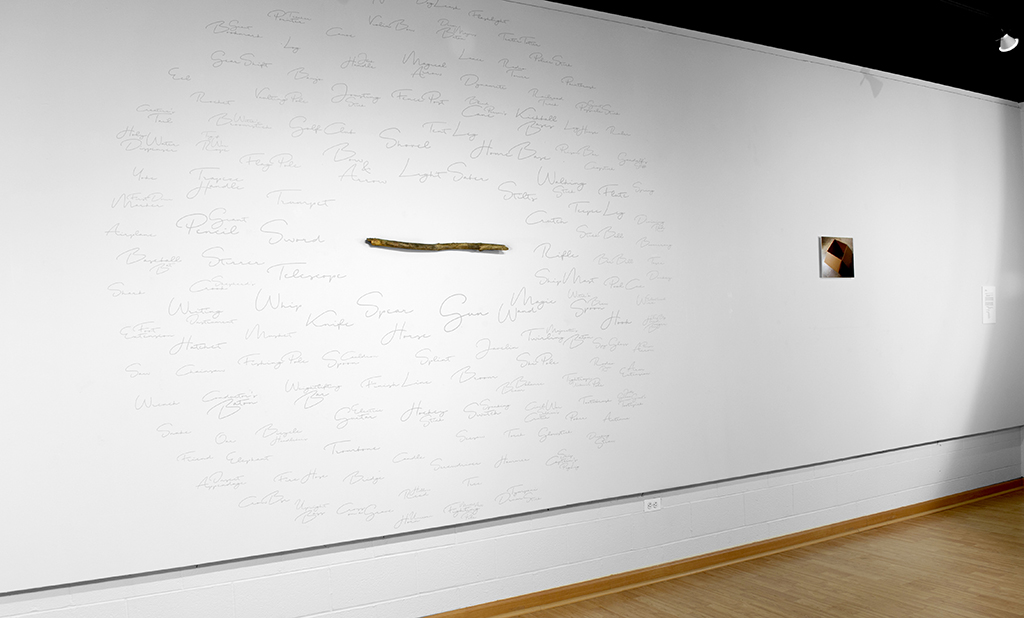 Installation showing the entire wall including the mounted stick with applied vinyl lettering and the photograph of the cardboard box and description of the piece.
